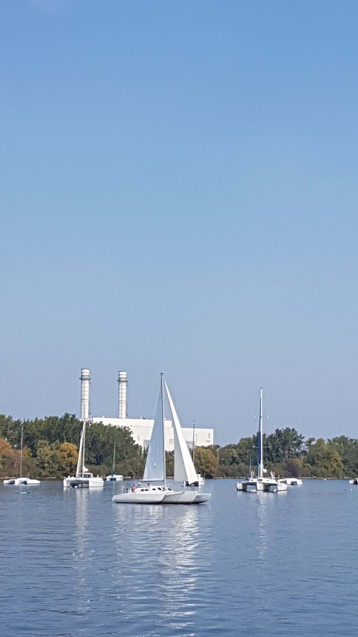 Sailboats on a calm lake with a blue sky and distant industrial buildings.