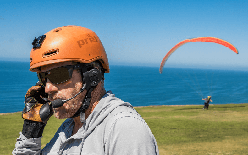 Man wearing MESH Communication Headset talking on the phone with a paraglider in the background.