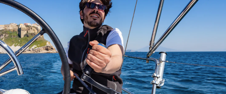 Man using a folding winch handle EasySea on a sailboat in the ocean with seaside scenery.